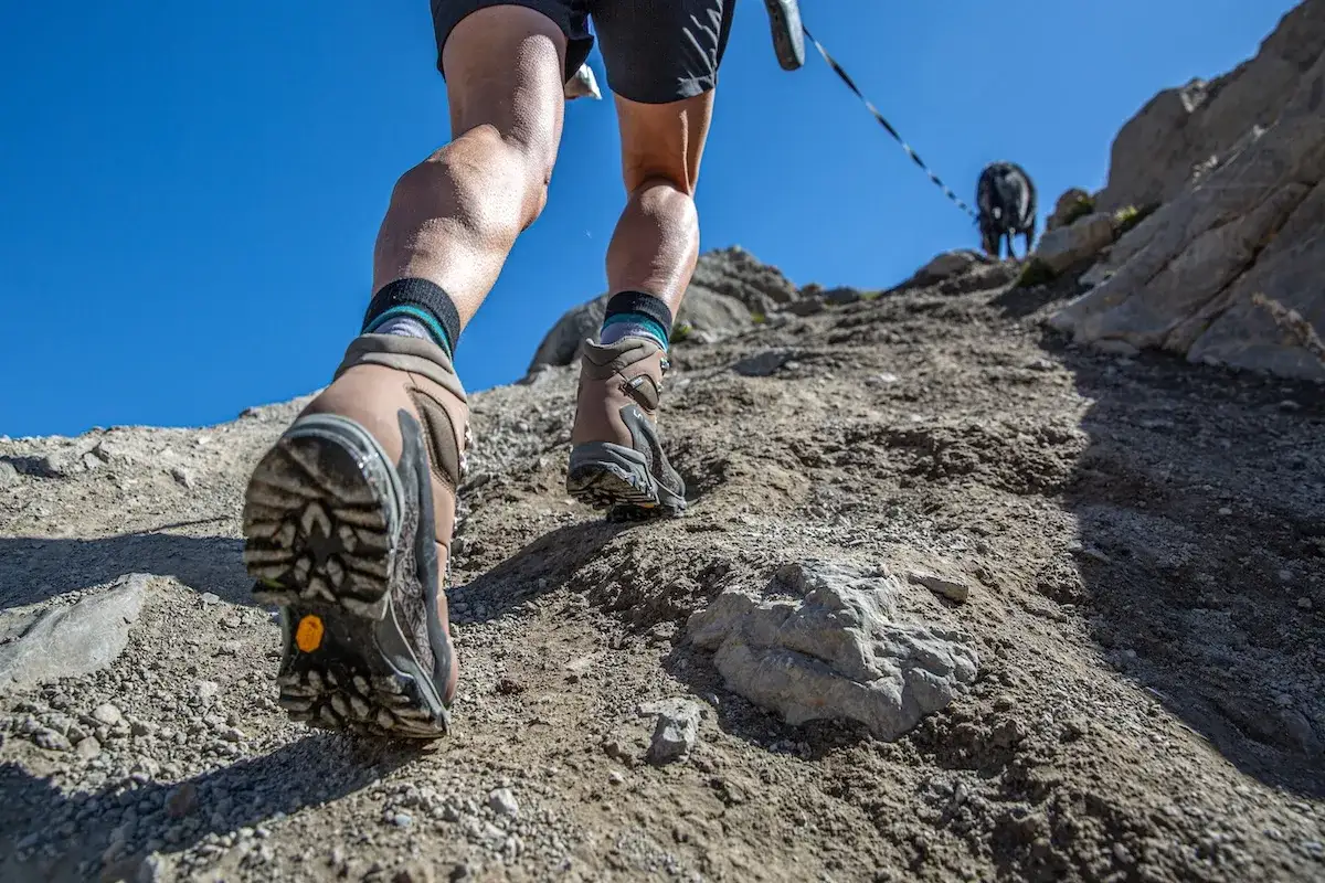 Choosing hiking boots is key for this rocky ascent. A person and their dog climb a steep, dusty trail under a clear blue sky.