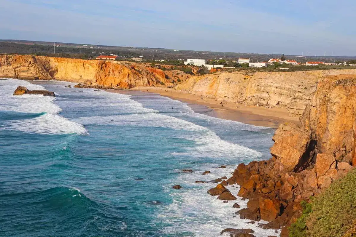 Waves crash on a sandy beach along the fisherman's trail in Portugal, with dramatic orange cliffs overlooking the turquoise ocean.
