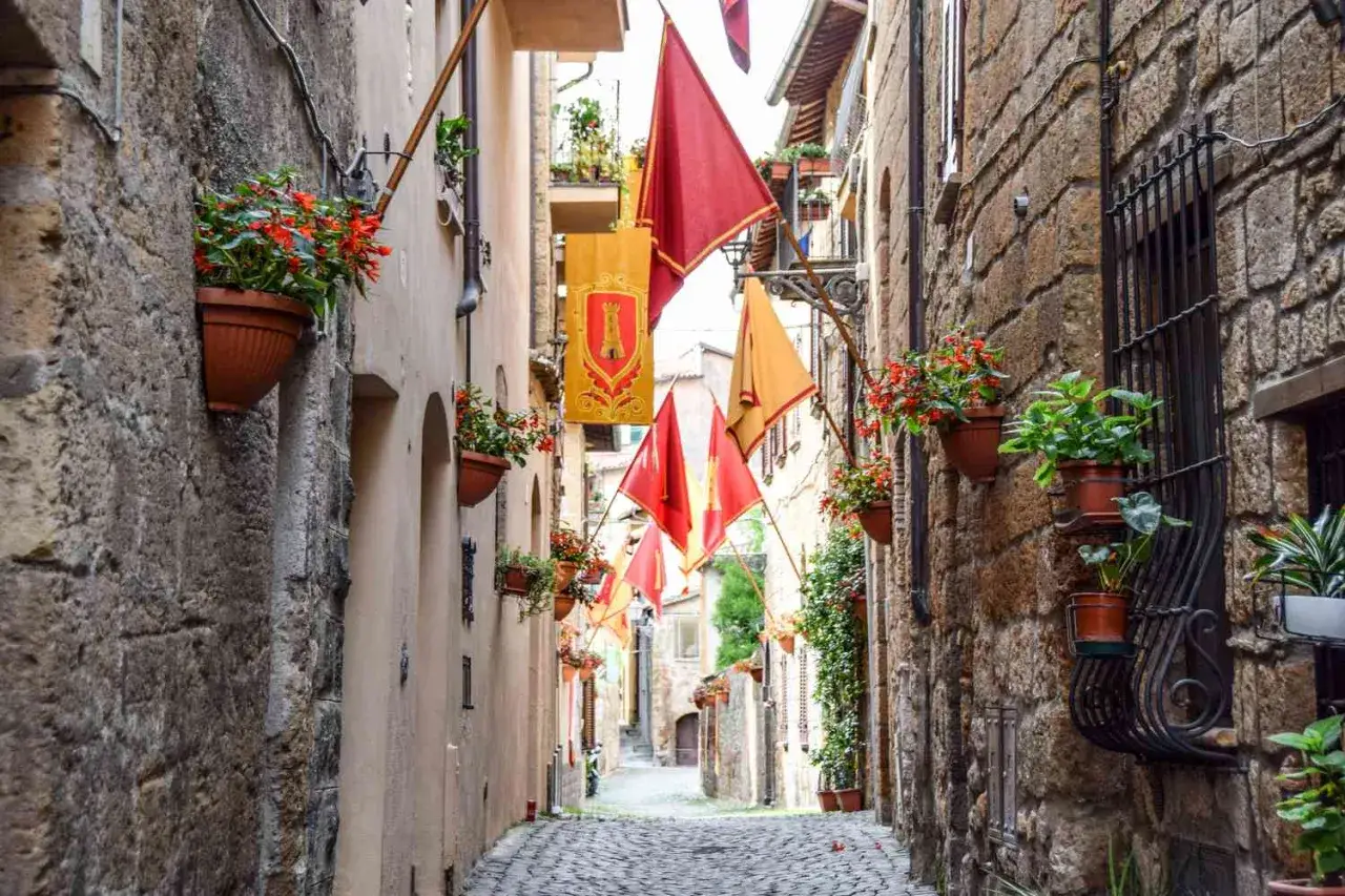 A charming cobblestone alley in Orvieto, adorned with red and gold flags and overflowing flower pots, beckons travelers on their journey to Rome.