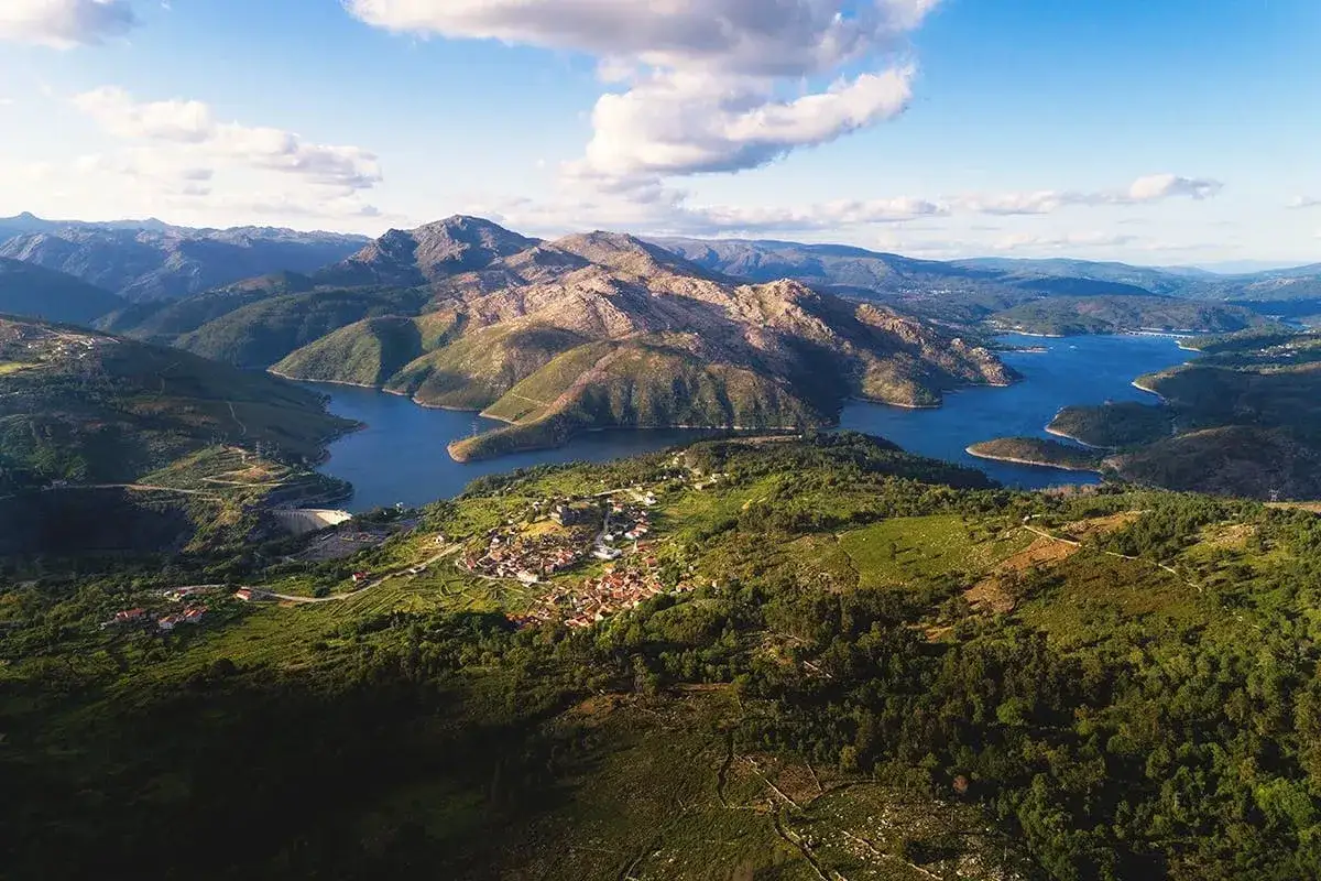 A scenic trail winds through a lush green landscape in Portugal, leading to a village nestled beside a serene blue lake and majestic mountains.
