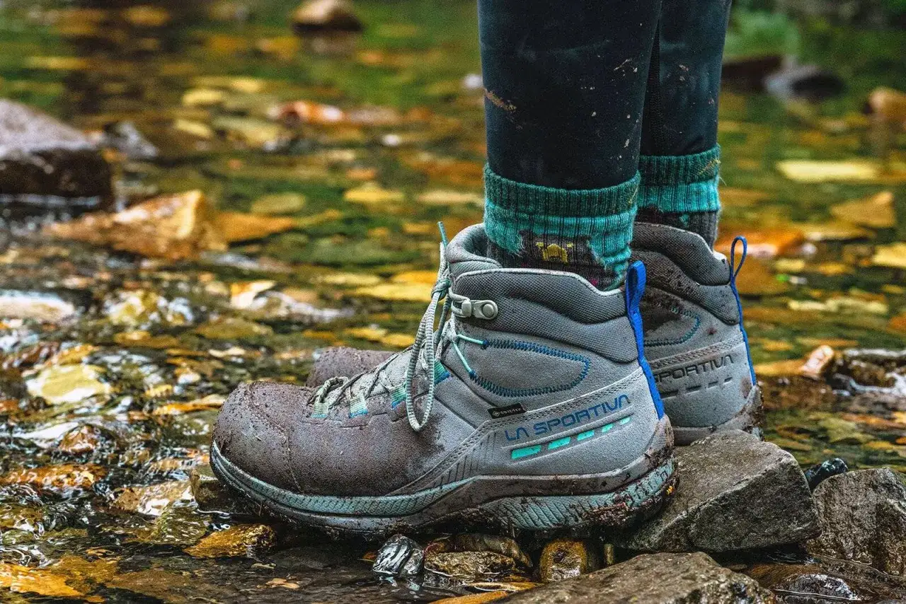 A hiker stands in a stream, testing the grip of synthetic hiking boots. The boots are muddy, showing they're ready for any terrain.