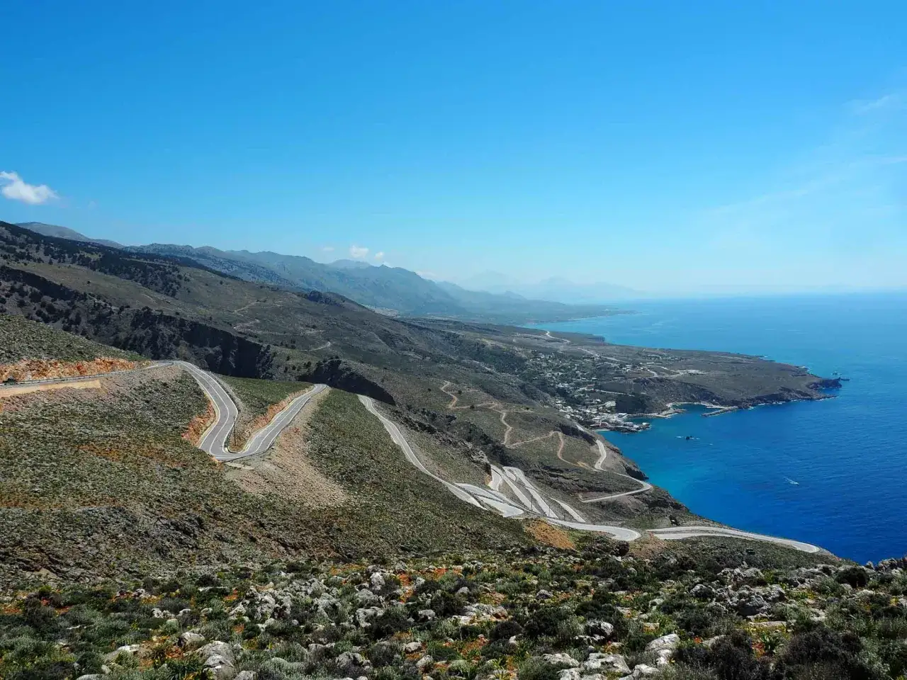 A winding mountain road snakes down to a coastal village in Greece. Is it easy to drive in Greece? This view suggests it can be challenging but rewarding!
