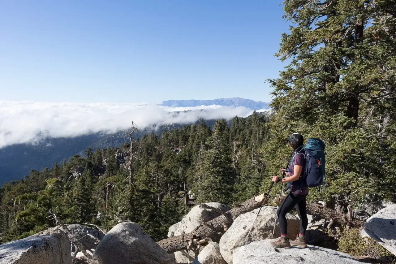 A woman in trail runners for backpacking pauses on a rocky overlook, gazing at a vast mountain landscape shrouded in clouds.