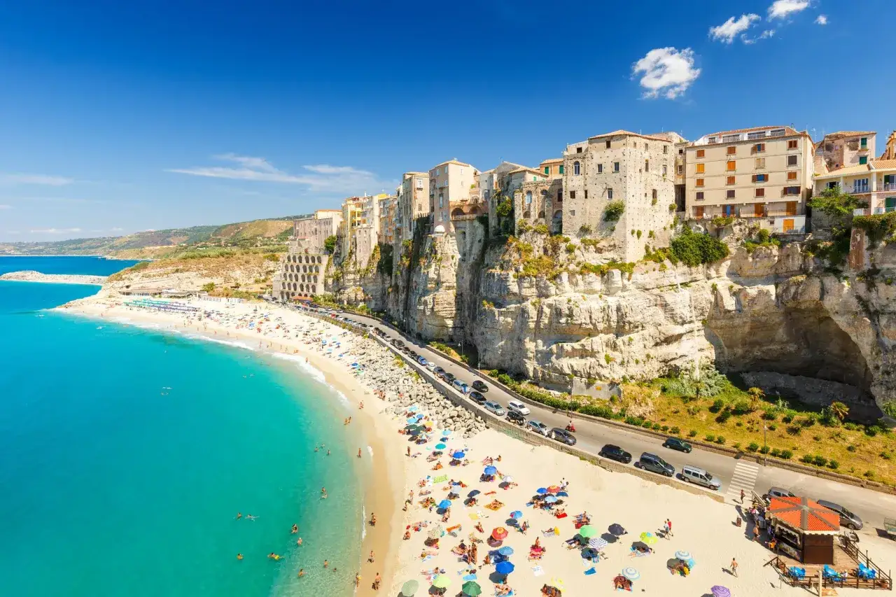 A vibrant beach scene in Calabria, Italy, with colorful umbrellas and people sunbathing. The town of Tropea clings to cliffs overlooking the turquoise sea, a must-see in Calabria.