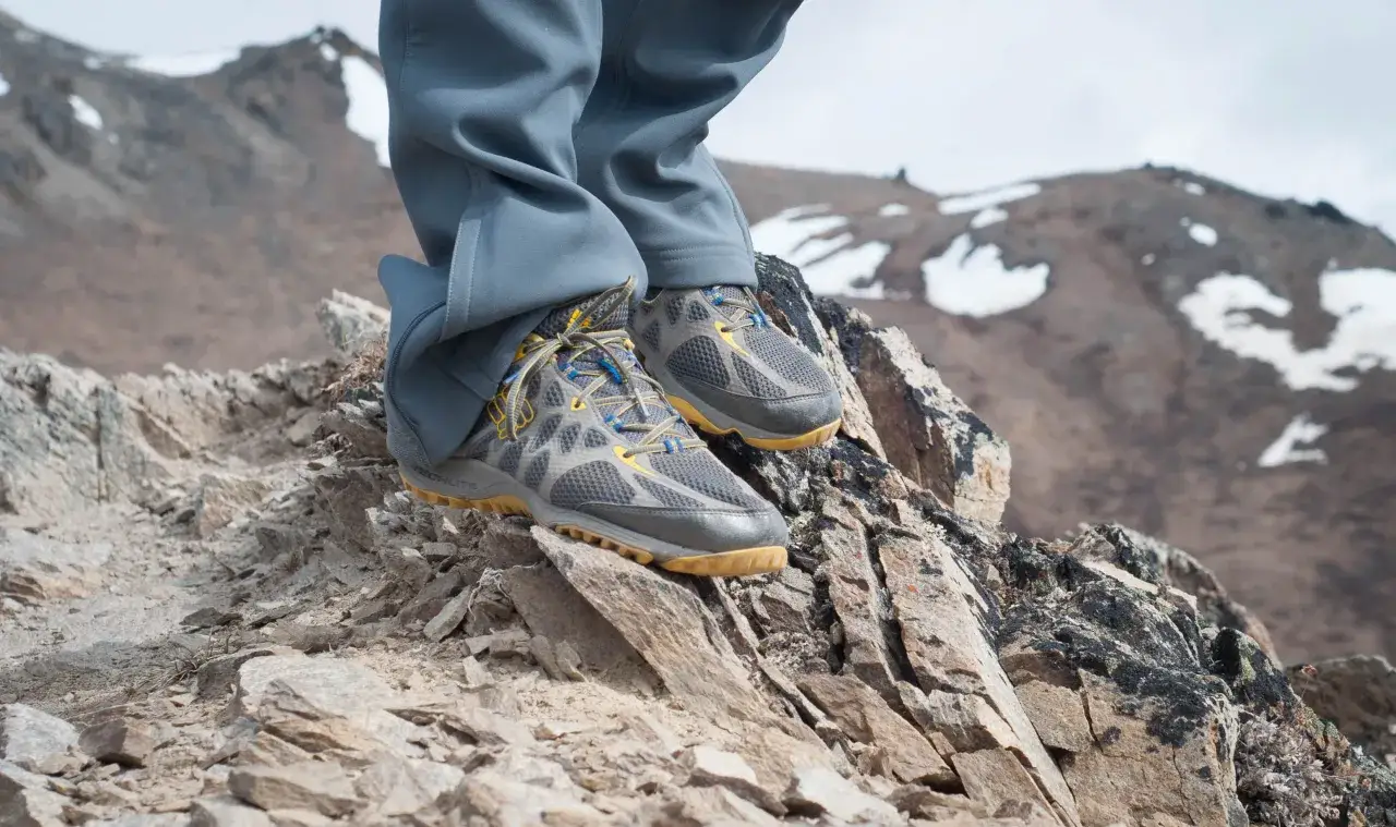 A hiker stands on a rocky mountain peak, wearing grey pants and Columbia OutDry hiking shoes, ready for adventure.