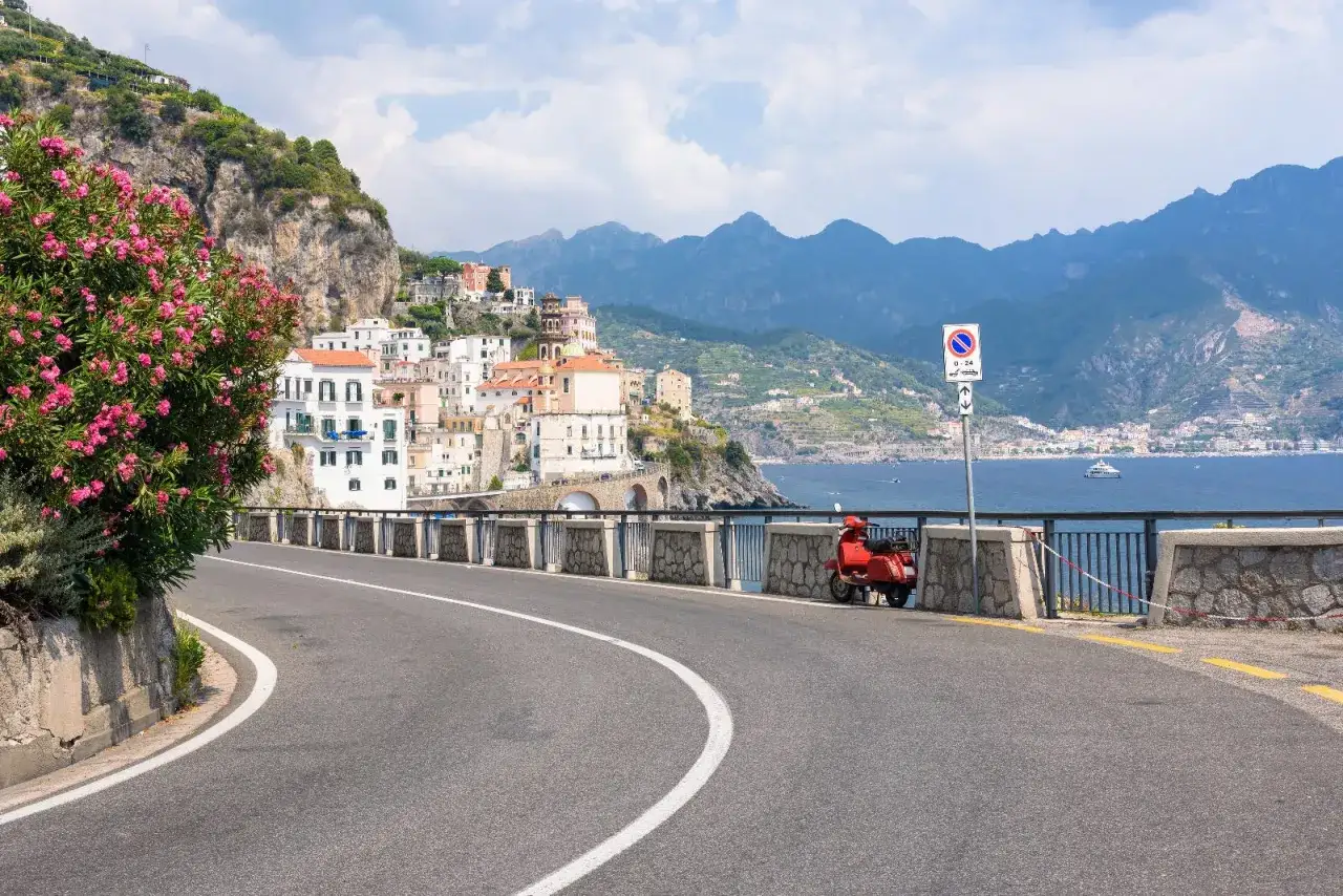 A red scooter waits on the scenic Amalfi Drive, overlooking a coastal town and the blue sea.