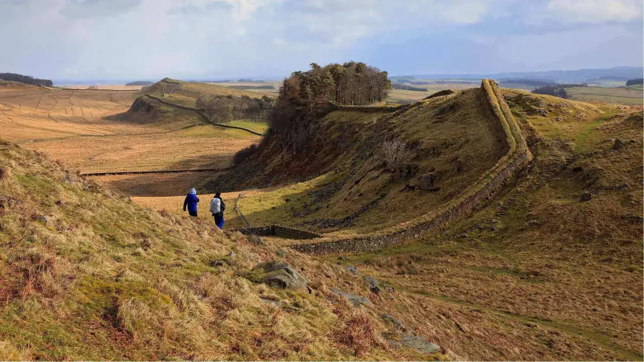 Hikers traverse a grassy ridge along Hadrian's Wall, a historic route reminiscent of the Pennine Way, with rolling hills and ancient stone walls stretching into the distance.