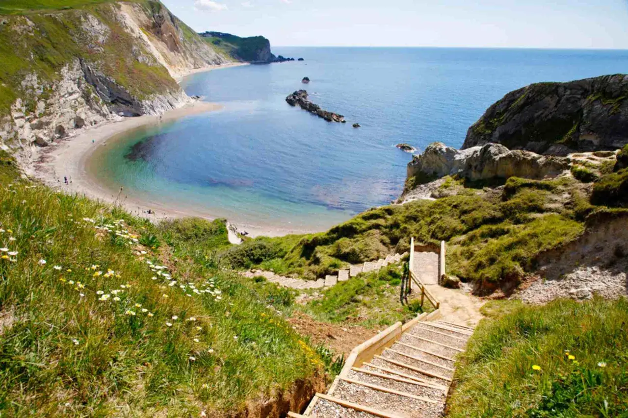 A wooden staircase descends along the salt path, leading to a secluded cove with turquoise waters and a sandy beach.