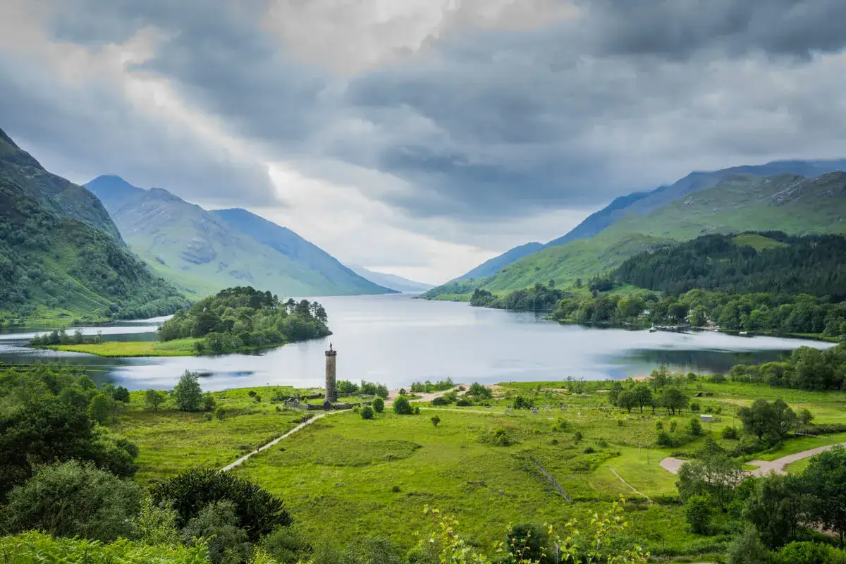 A dramatic view of a Scottish loch, perfect for Scotland hiking trips. Mountains rise on either side, with a monument on a small island.