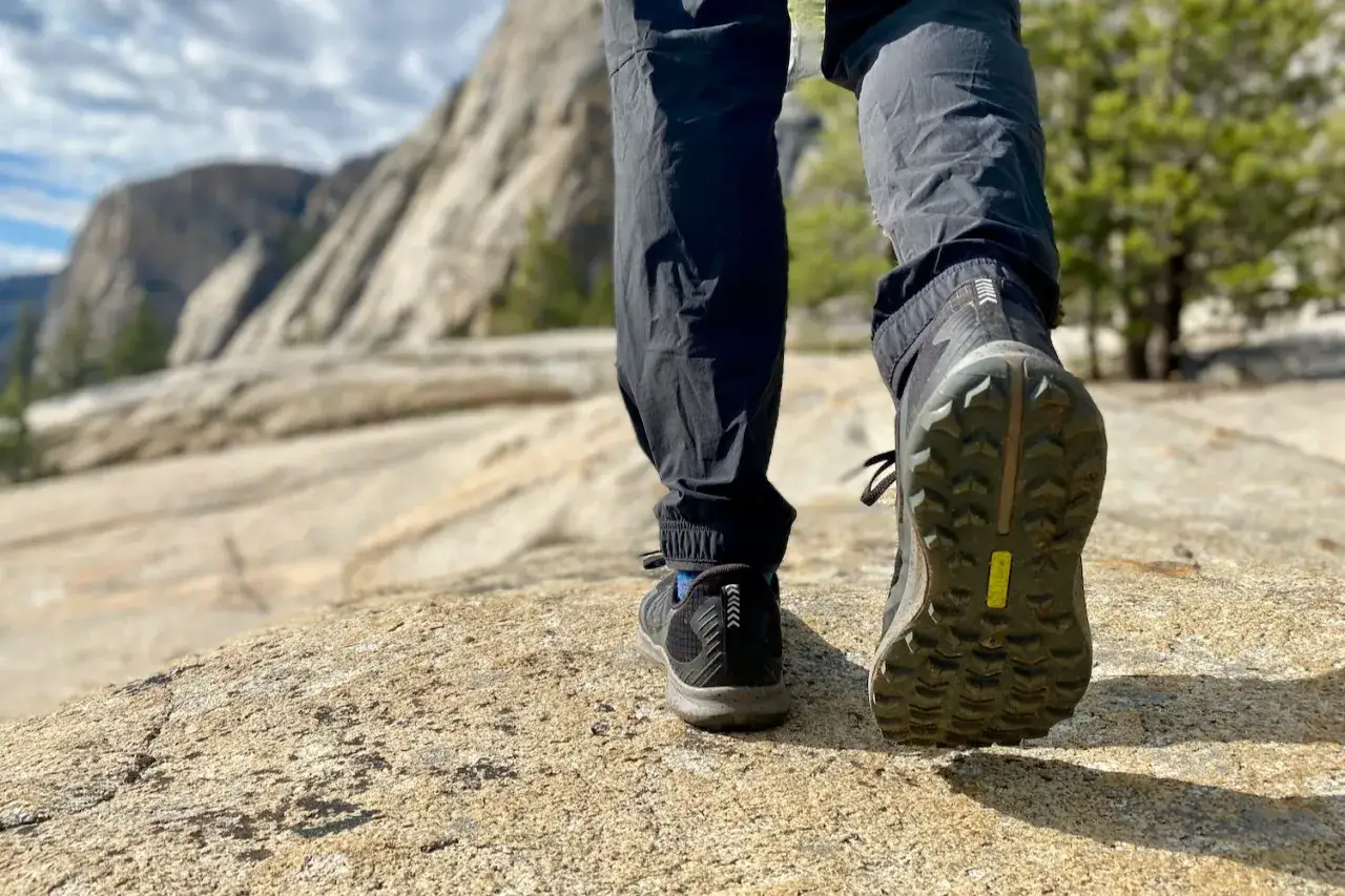 A hiker's feet, wearing shoes with a deep lug sole for grip, ascend a rocky mountain trail. The lug sole means the tread has deep, prominent patterns for traction.