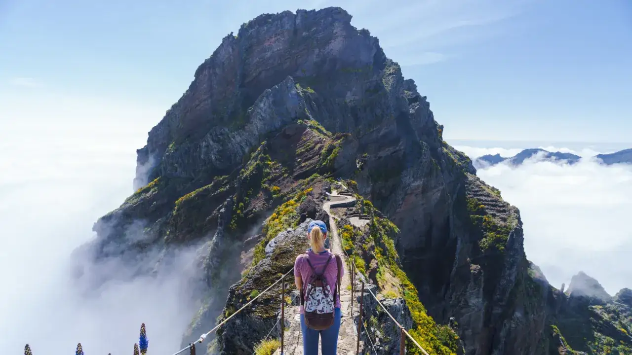 A hiker stands on a narrow path, ready for long distance hikes in Europe. The dramatic mountain peak looms ahead, with clouds swirling below.