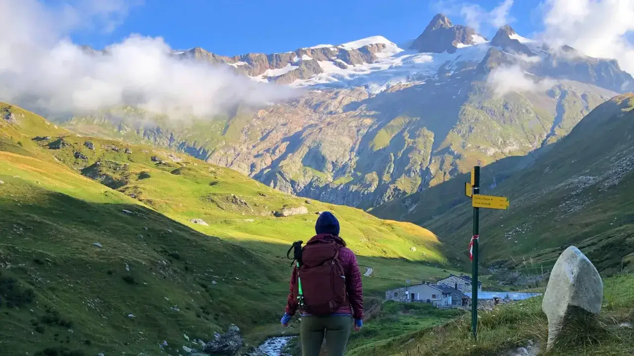 A hiker pauses on a Tour Mont Blanc hike, gazing at a majestic snow-capped mountain range. A signpost points the way to mountain huts.