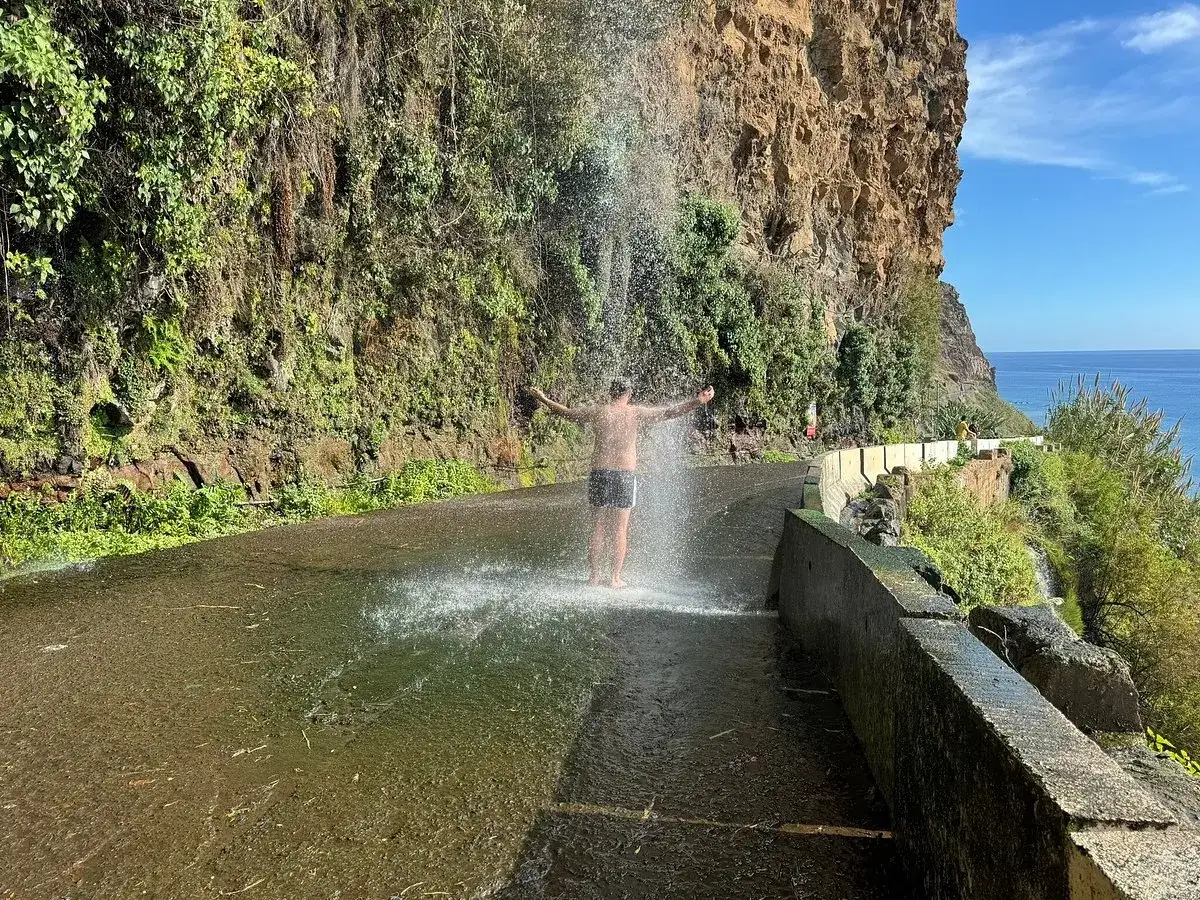 A refreshing moment during a Madeira hike, a person stands under a waterfall cascading onto a road beside a cliff.