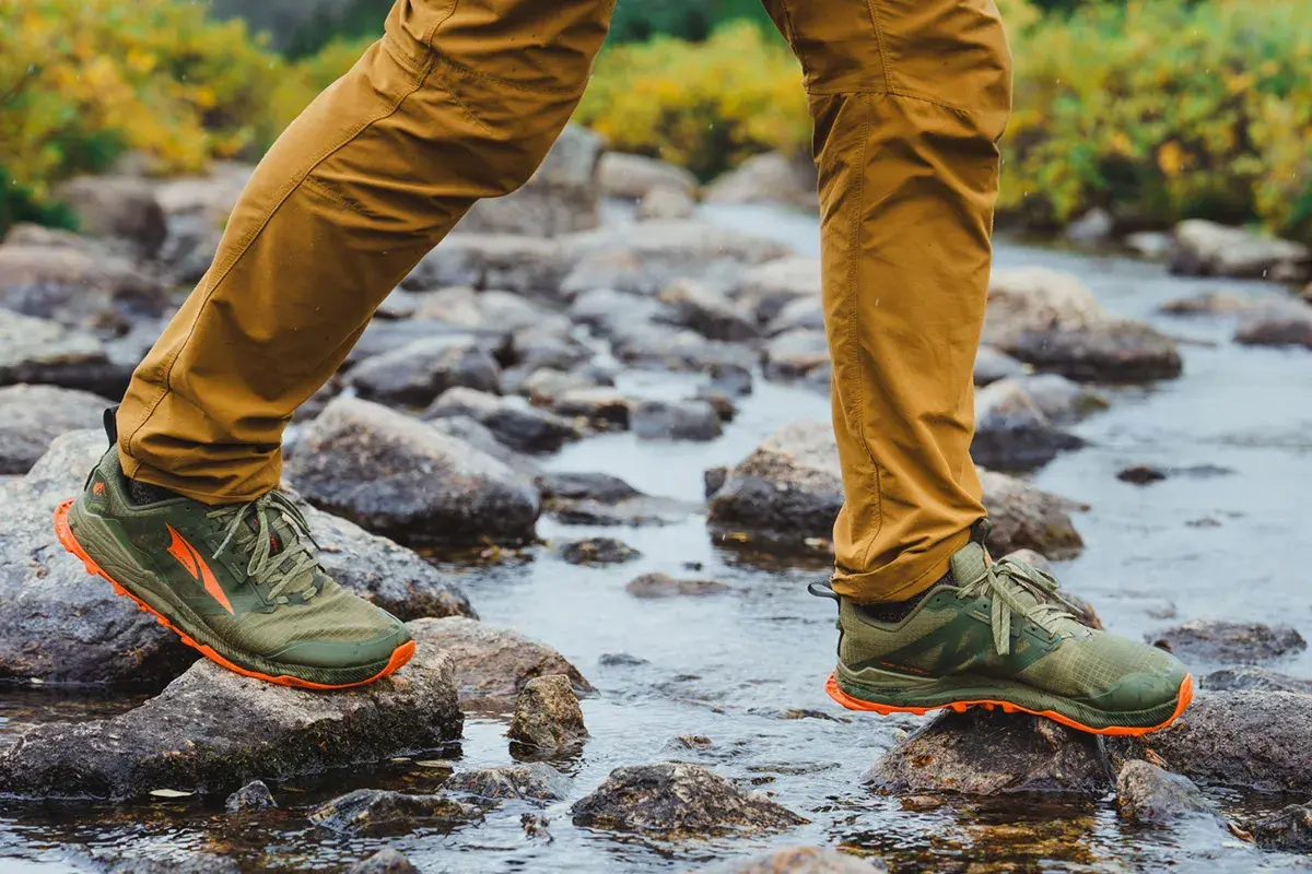 A person walks across a rocky stream in olive green hiking shoes with orange accents. These shoes are built for adventure, but can you wear hiking shoes everyday?