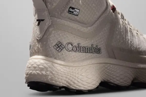 Close-up of a beige Columbia hiking boot with water droplets. The boot features the Columbia logo and "OutDry WATERPROOF" branding, showcasing its durable, water-resistant design.